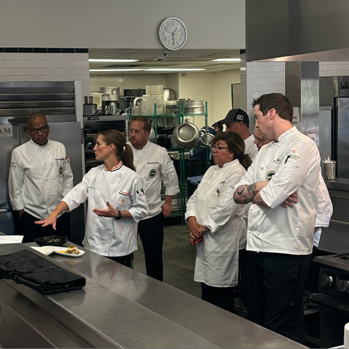 A group gathered around a chef who is explaining something